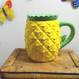 May include: A yellow pineapple-shaped mug with a green handle and lid, sitting on a wooden surface. The mug has a textured surface and is decorated with small black dots. Colorful butterfly decorations are on the wall behind the mug.