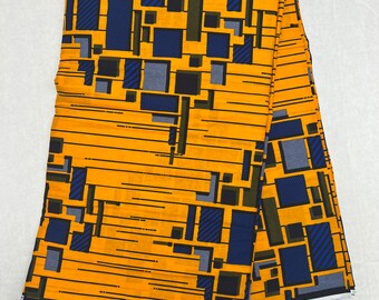 May include: Folded fabric with a geometric pattern. The fabric has a mustard yellow background with a design of blue, black, and grey rectangles and lines. The word "VERITA" is visible on the edge of the fabric.