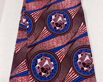 May include: Folded fabric with a striking geometric design. The pattern includes repeating circular motifs with blue and purple details, set against a red and black striped backdrop. The fabric has a label that reads "AX BLOCK PRINTS HITA".