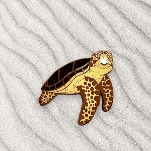 Puede incluir: Un pin de tortuga marina marrón y amarillo con un diseño detallado. La tortuga está mirando hacia arriba con la cabeza levantada y la boca ligeramente abierta.