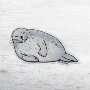 Puede incluir: Una foca gris con una expresión feliz, acostada boca abajo con las aletas metidas debajo del cuerpo. La foca tiene un patrón de manchas y está dibujada en un estilo de dibujos animados. La imagen tiene un fondo blanco y el texto "@FinPinShop" es visible en la esquina inferior derecha.