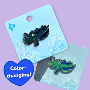 May include: Two color-changing enamel pins featuring a cuttlefish design. The pins are on blue and white card stock with a white outline of seaweed and coral. The text "Fin Pin" is in the top left corner of the card stock. The text "Color-changing!" is in a blue heart shape.
