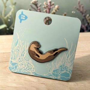 May include: A brown enamel pin shaped like a swimming otter attached to a light blue backing card. The card features white line art of coral and seaweed, with the text FIN PIN printed in the top left corner and a circular hole at the top.