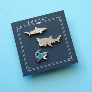 Könnte beinhalten: Drei Emaille-Pins mit verschiedenen Haiarten. Die Pins sind silberfarben mit blauen und weißen Details. Die Pins werden auf einem dunkelblauen Hintergrund mit dem Wort "SHARKS" in weißen Buchstaben angezeigt.