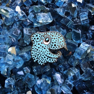 May include: A blue and black enamel pin of a cartoon squid with a large eye and a spotted pattern.
