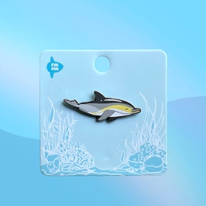 Peut inclure: Une épingle en émail bleu et blanc représentant un dauphin de dessin animé avec une bande jaune sur le côté. L'épingle est sur une carte blanche avec une bordure bleue et le texte "FIN PIN" dans un cercle bleu.