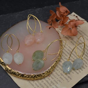 Op de afbeelding: Goudkleurige oorhangers met druppelvormige edelstenen in verschillende kleuren, waaronder roze, groen en wit. De oorbellen worden gepresenteerd op een decoratief dienblad met een gouden rand.