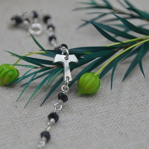 Pode incluir: Um bracelete de prata com um fecho de cruz de prata e contas pretas. O bracelete está sobre uma superfície de tecido bege com folhas verdes e vagens de sementes verdes.