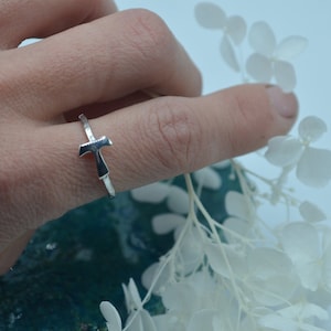 Könnte beinhalten: Ein silberner Ring mit einem schlichten, gehämmerten Kreuzdesign. Der Ring wird an einem Finger getragen und ist von weißen Blumen umgeben.