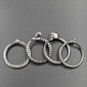 May include: Four silver rings in a row, each with a different design. The first ring is a snake eating its tail, the second is a snake with a bird head, the third is a snake with a skull head, and the fourth is a snake eating its tail.