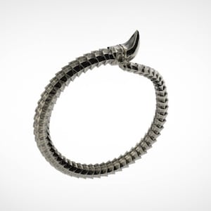 May include: A silver-toned, articulated bracelet with a segmented design, resembling a stylized snake or dragon. The bracelet curves in a circular shape, with a pointed, horn-like element at one end. The background is plain white.