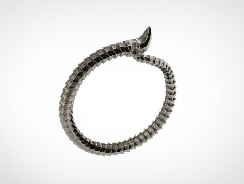 May include: A silver-toned, articulated bracelet with a segmented design, resembling a stylized snake or dragon. The bracelet curves in a circular shape, with a pointed, horn-like element at one end. The background is plain white.