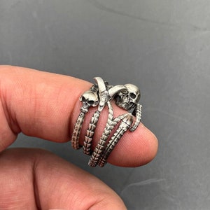 May include: A silver-toned ring featuring three skull designs and a skeletal claw motif. The ring has a detailed, gothic aesthetic, with the skulls and claw elements intricately crafted. The ring is held up by a finger.