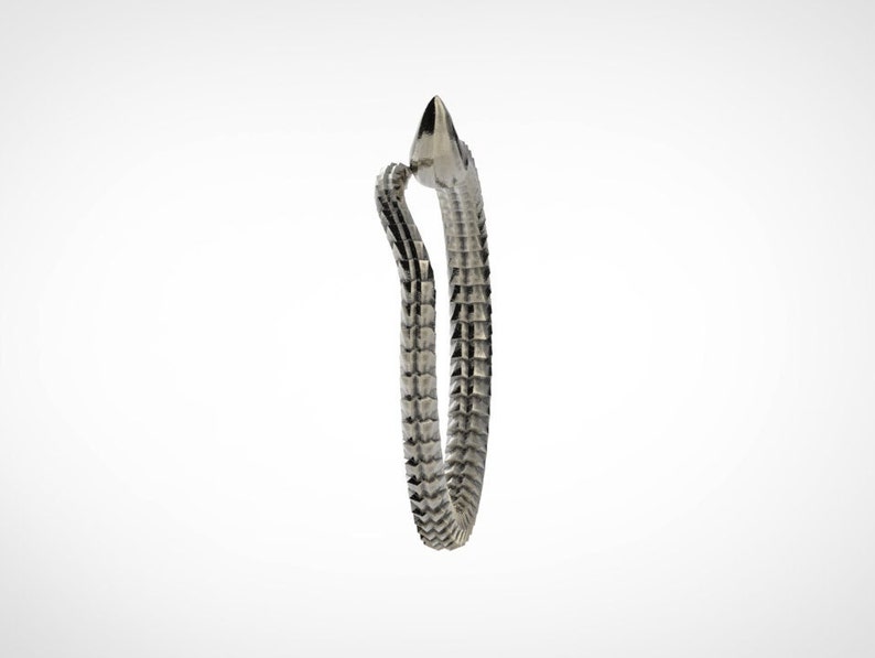 May include: A silver-colored bracelet with a crocodile design. The bracelet features a textured, scale-like pattern along its band, and a crocodile head with a pointed snout. The bracelet is set against a white background.