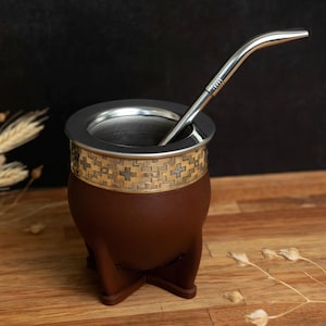 Puede incluir: Una calabaza de yerba mate marrón con una banda dorada decorativa con un patrón de cruces. Tiene un borde de acero inoxidable y una pajita bombilla a juego. La calabaza se asienta sobre una base de tres patas, ideal para bebidas sudamericanas.