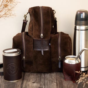 May include: A collection of brown suede travel accessories, including a backpack, a thermos, a tea canister, and a leather cup with a metal straw. The items are arranged on a wooden surface.