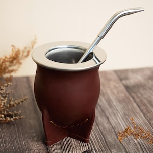 Puede incluir: Una taza de mate de cuero marrón con borde y base de acero inoxidable, y una pajita de acero inoxidable. La taza tiene una base única de tres patas y está diseñada para beber té tradicional sudamericano.