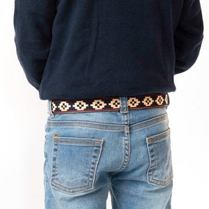 May include: A person wearing blue jeans with a woven belt featuring a geometric pattern in red, black, and cream.