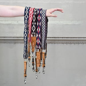 Puede incluir: Un conjunto de siete correas para cámara con diferentes correas tejidas con patrones y detalles en cuero marrón. Las correas cuelgan del brazo de una persona.