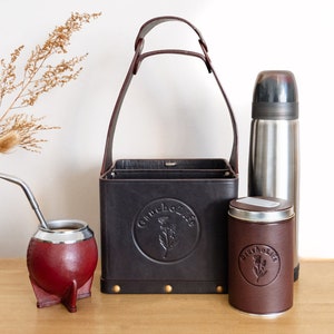May include: A collection of items for enjoying yerba mate. Includes a dark brown leather carrier, a stainless steel thermos, a red gourd with a metal straw, and a tin of tea. The carrier and tin have the "Gaucho Life" logo.