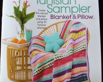 May include: A crochet pattern book featuring a colourful striped blanket and pillow. The book cover features the title "Tunisian Sampler Blanket & Pillow" and the text "Create this vibrant blanket and pillow using 33 Tunisian stitches!"