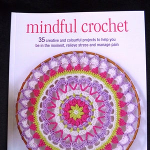 May include: A book titled "mindful crochet" with a colourful crochet mandala design. The book cover features a wooden embroidery hoop with a vibrant crochet pattern in shades of pink, purple, green, and white. The text reads "35 creative and colourful projects".