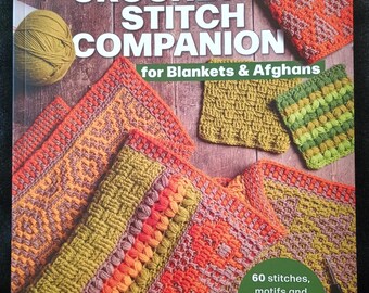May include: A copy of "The Crochet Stitch Companion" by Anna Nikipirowicz, showcasing crochet stitch examples for blankets and afghans. The cover features the title, author, and crochet patterns in orange, green, and yellow. Includes text: "60 stitches, motifs and edges to create your perfect project."