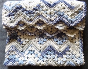 May include: A folded, handmade crochet blanket in shades of blue, white, and gray. The blanket features a detailed pattern with a scalloped edge and geometric designs. The soft, textured blanket is perfect for a baby or as a decorative throw.