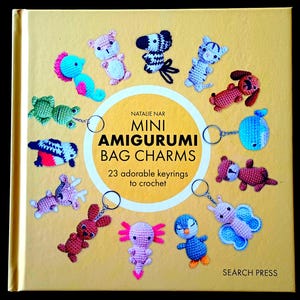 Op de afbeelding: Een vierkant boek met een gele kaft getiteld Mini Amigurumi Bag Charms: 23 adorable keyrings to crochet door Natalie Nar. De kaft toont diverse kleurrijke gehaakte dierenfiguren in een cirkel, waarvan sommige zijn bevestigd aan metalen sleutelringen.
