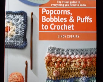 May include: Book cover with the title "Popcorns, Bobbles & Puffs to Crochet" by Lindy Zubairy. The cover features colorful crocheted samples and crochet hooks.
