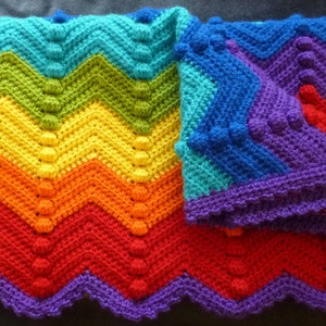May include: A handmade crochet blanket in a vibrant rainbow of colors, including red, orange, yellow, green, blue, and purple. The blanket features a chevron pattern with textured bobbles, and a scalloped purple border. A cozy, colorful home decor item.