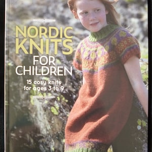 May include: Book cover featuring a young girl wearing a red, green, and yellow knitted dress with a Nordic pattern. The title of the book is "Nordic Knits for Children" and the subtitle is "15 cosy knits for ages 3 to 9".