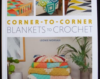 May include: Book cover featuring the title "Corner-to-Corner Blankets to Crochet" by Leonie Morgan. The cover shows three colourful crocheted blankets in different patterns and colours, displayed on a bed, a chair, and a cot.