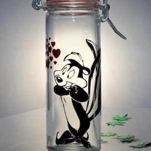 May include: A clear glass jar with a metal lid and a cartoon image of a skunk with red hearts. The jar is on a white surface with green leaves scattered around it.