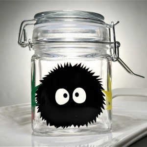 May include: A clear glass jar with a metal lid and a black and white cartoon character on the front. The character has large eyes and a fuzzy appearance. The jar is decorated with green, yellow, and red accents.