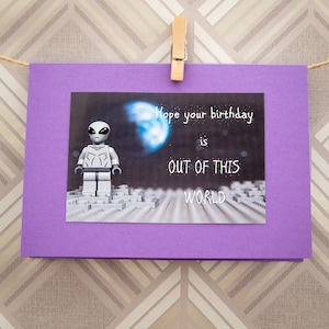 May include: A purple card with a picture of a gray alien Lego minifigure standing on a gray and white Lego baseplate. The background is a dark space scene with a blue planet and stars. The text on the card reads "Hope your birthday is out of this world."