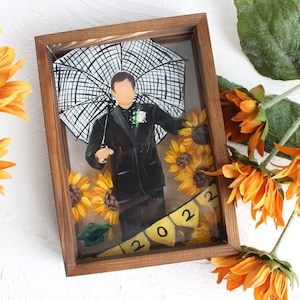 Puede incluir: Una obra de arte enmarcada que presenta a una persona sosteniendo un paraguas, rodeada de girasoles y una pancarta con el año 2022.