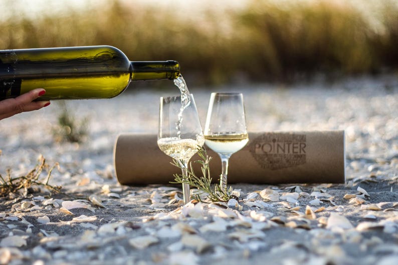 2 picnic white wine glasses pinned to the sand on the beach with white wine bottle and white wine being poured in them