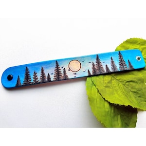 May include: A blue leather bracelet with a painted design of a forest scene with a full moon and birds flying overhead.