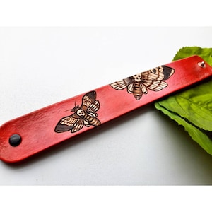 May include: Red leather bracelet with two engraved images of moths. The moths have a skull and crossbones design.