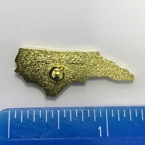 Puede incluir: Pin de metal de color dorado con forma del estado de Carolina del Norte. La parte posterior del pin tiene un peque&ntilde;o poste de metal para sujetarlo a la ropa o a una bolsa. El pin tiene el texto "es@stl76.com" en la parte posterior.