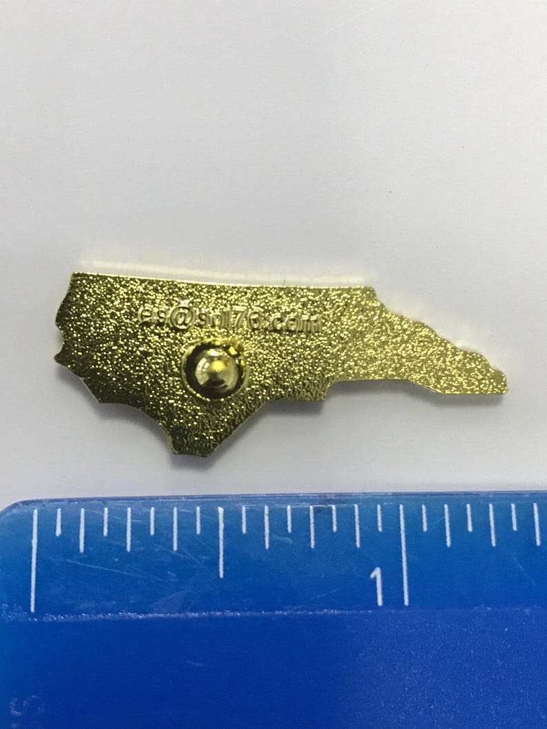 Puede incluir: Pin de metal de color dorado con forma del estado de Carolina del Norte. La parte posterior del pin tiene un peque&ntilde;o poste de metal para sujetarlo a la ropa o a una bolsa. El pin tiene el texto "es@stl76.com" en la parte posterior.
