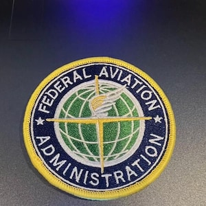 May include: A blue and yellow embroidered patch with the text "Federal Aviation Administration" and a globe with a wing design.