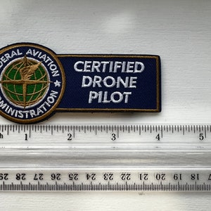 May include: A blue and gold embroidered patch with the text "CERTIFIED DRONE PILOT" and the Federal Aviation Administration logo. The logo features a globe with a lightning bolt and the words "FEDERAL AVIATION ADMINISTRATION".