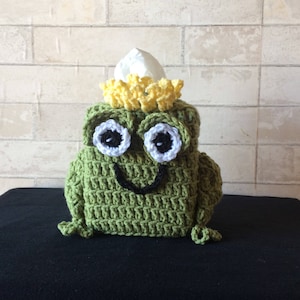 May include: Green crocheted frog tissue box cover with a yellow crocheted crown on top. The frog has white eyes with black pupils and a black smiling mouth.