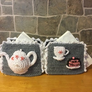 May include: Two crocheted napkin holders, one with a teapot and the other with a teacup and a cake. Both holders are gray with white crocheted trim and feature pink accents.
