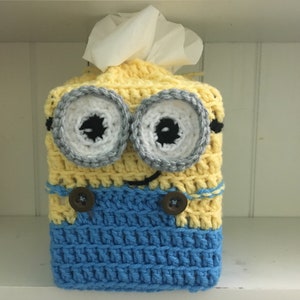 May include: A handmade crochet tissue box cover in the shape of a Minion character. The cover is primarily yellow and blue, with large, round eyes, and a smiling mouth. The tissue box cover has two brown buttons and white tissues are coming out of the top.
