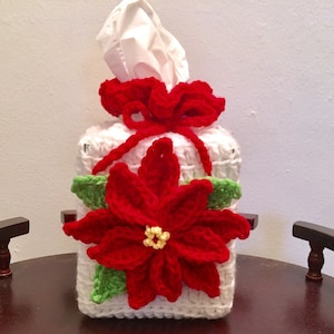 May include: White crocheted tissue box cover with a red poinsettia flower and green leaves. The cover has a red crocheted bow on top.