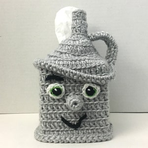 May include: A gray crocheted tissue box cover shaped like a jug with a smiling face, green eyes, and a black nose.