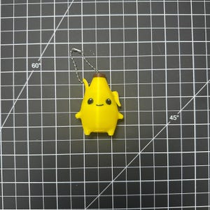 May include: A yellow banana-shaped keychain with a brown top, featuring a printed face with two black eyes and a small smile. The figure has small arms and legs, and is attached to a silver-colored metal ball chain for hanging purposes.